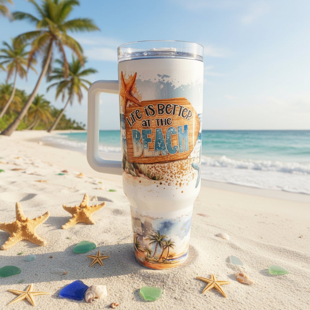A clear handled tumbler with a white handle and a tropical beach-themed wrap stands on a sandy beach. The wrap features a wooden sign reading 'Life is Better at the Beach' with starfish and palm tree illustrations.