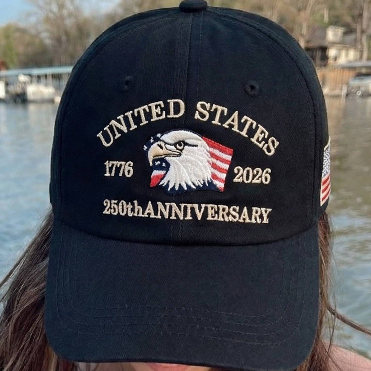 Black cap with 'United States 1776-2026 250th Anniversary' text and eagle design worn by a person by a body of water.