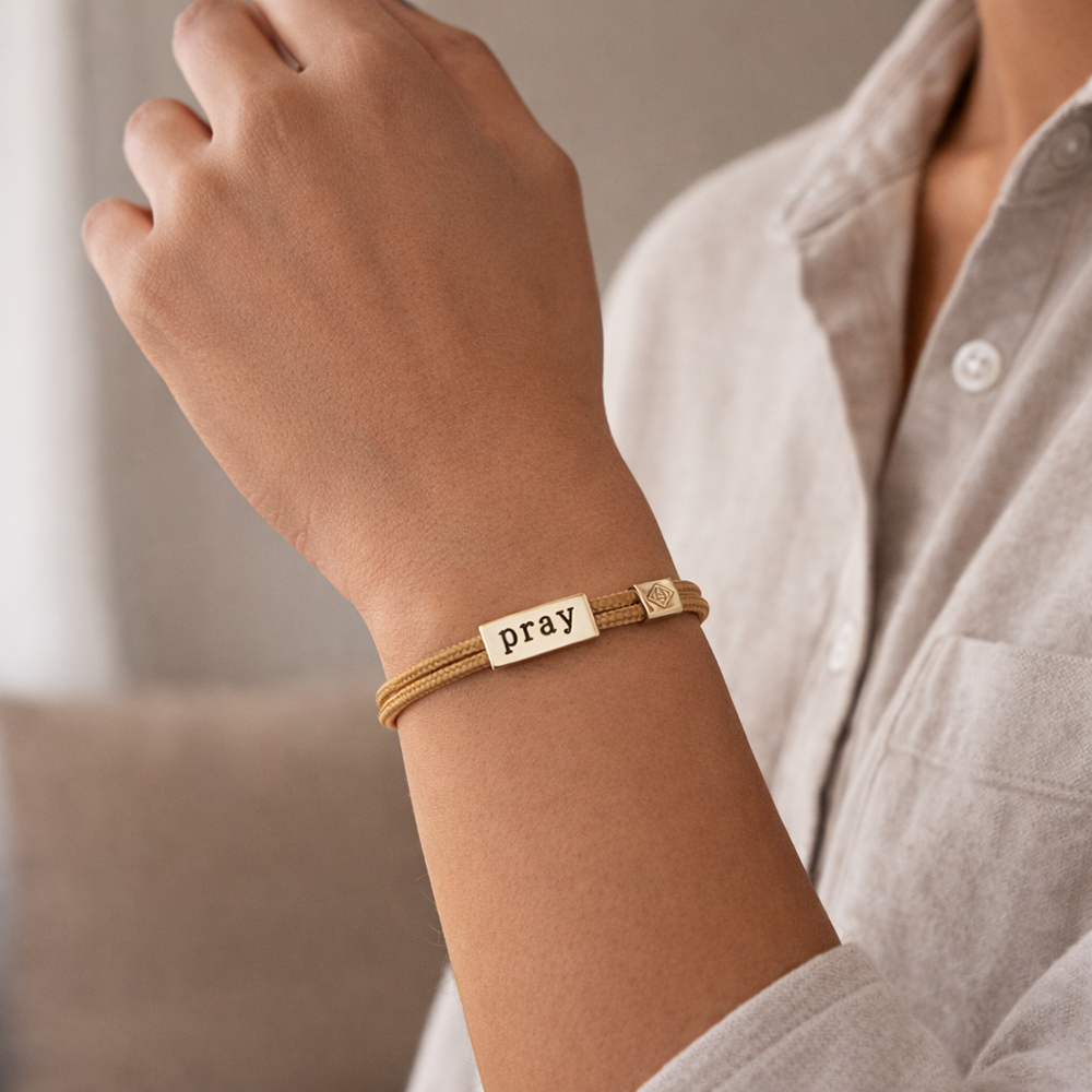 Gold bracelet with 'pray' tag on wrist against neutral background