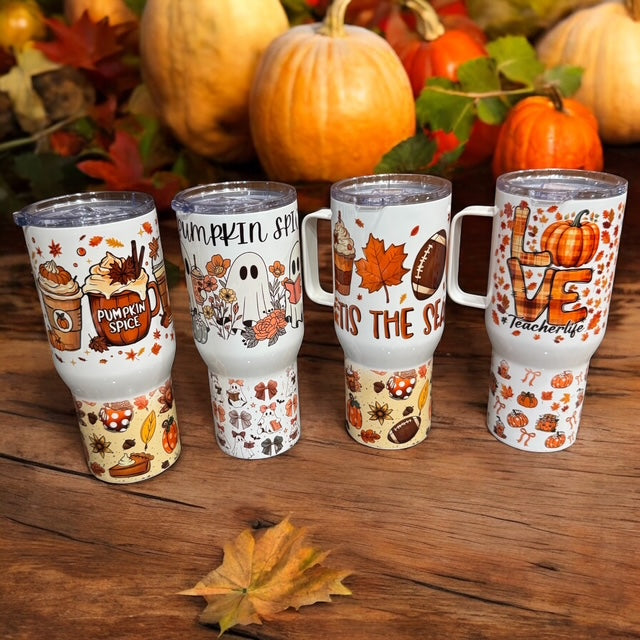 Four white handled tumblers with autumn-themed designs, including pumpkins, fall leaves, ghosts, and the phrase 'Pumpkin Spice Love is the best', arranged on a wooden surface with pumpkins and fall leaves in the background.
