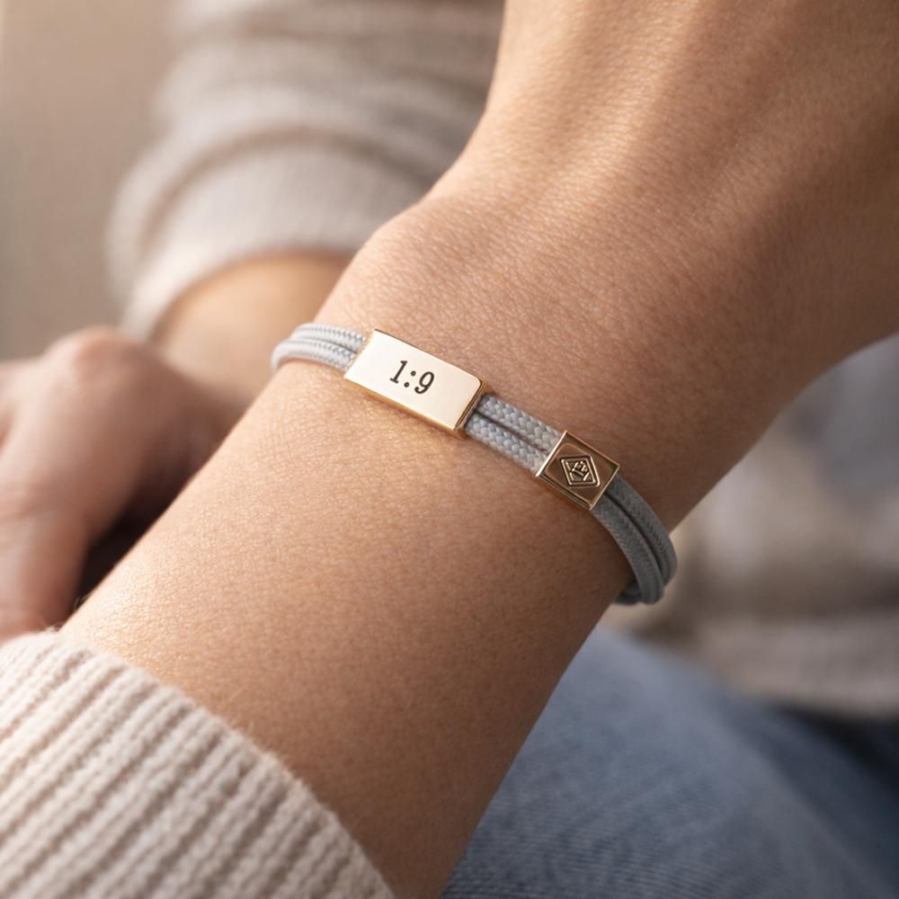 Bracelet with a gold tag and gray strap on a wrist, worn by a person.